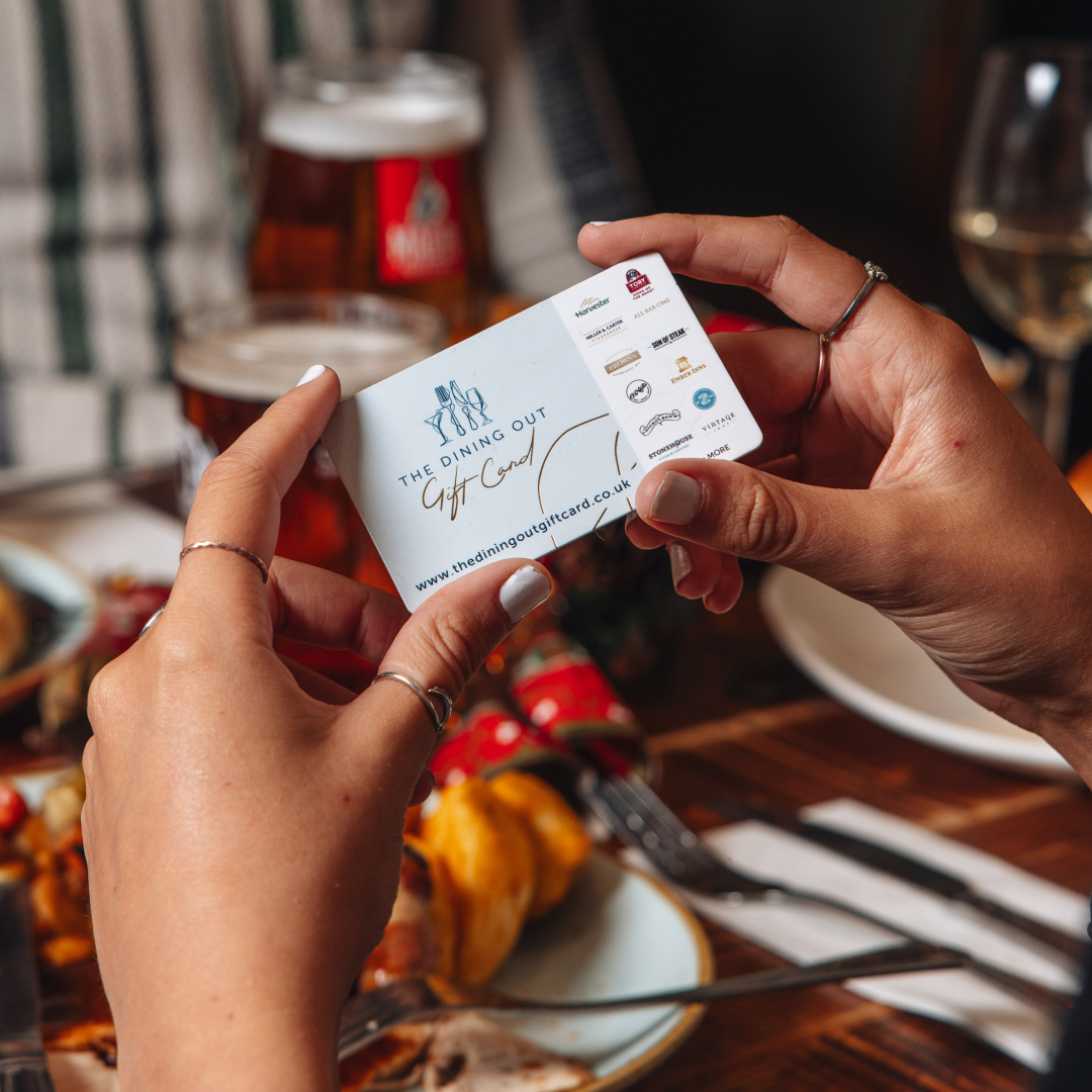 A person holding a physical Dining Out Gift Card in their hands. In the background is a table with drinks on it. 